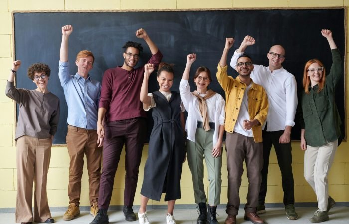 A group of eight adults cheerfully standing with raised fists in front of a blackboard indoors.