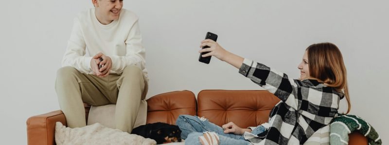 Happy teenagers using a smartphone for selfies while relaxing on a sofa indoors.
